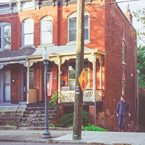 The image shows a man standing beside a red brick house. For accessibility purposes, a concise alt text could be: "Man standing beside a red brick townhouse."