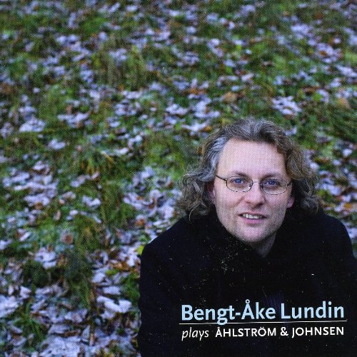 Man with curly hair and glasses smiles in a grassy area with fallen leaves.