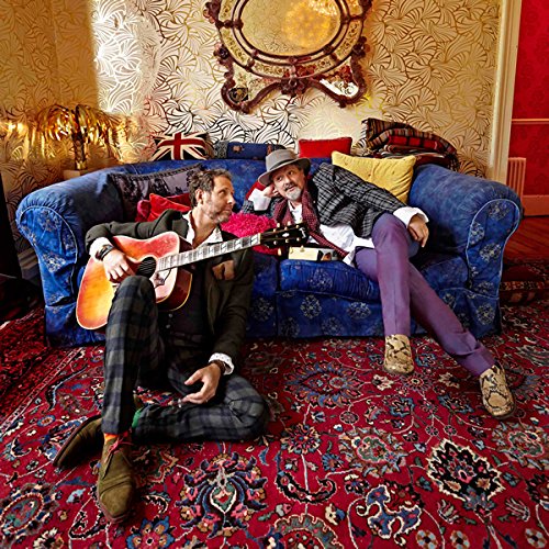 Two men, one with a guitar, relax on and beside a blue couch on a red rug.