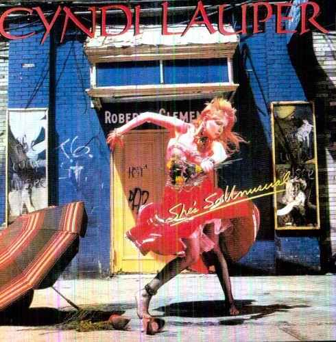"Album cover featuring Cyndi Lauper in a red dress dancing outside a blue building."