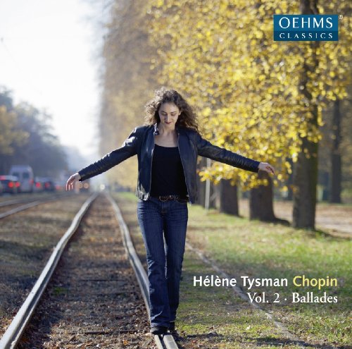 A woman stands on train tracks with trees and vehicles in the background.