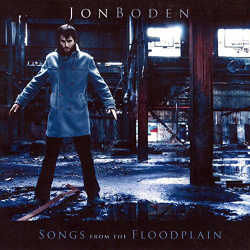 "Album cover featuring a man in a blue coat standing in a dimly lit, flooded industrial space."