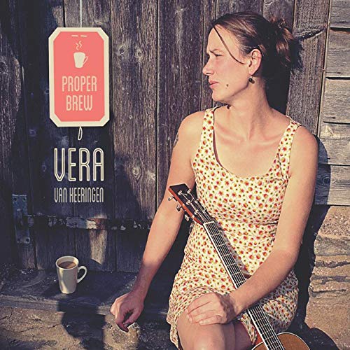 Woman in spotted dress holding guitar, beside a coffee cup, against a wooden wall.