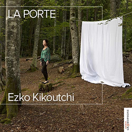 Woman stands on tree stump in forest, with white sheet hanging nearby.