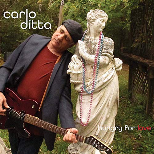 "Man in dark jacket holding guitar beside a beaded statue in a garden."