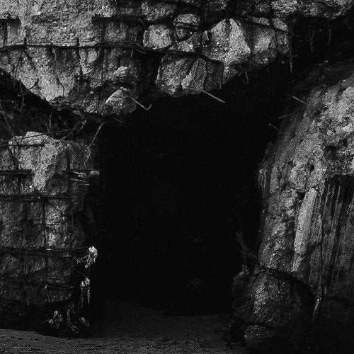 The image depicts a dark cave entrance surrounded by barbed wire and rocks. For accessibility purposes, a suitable alt text could be: "A black and white image of a cave entrance."