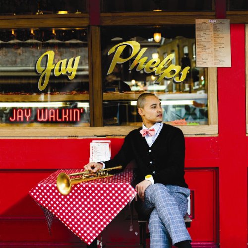 Man sits at small table outside red restaurant, holding trumpet, wearing bow tie and cardigan.