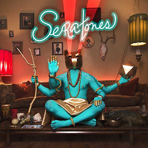Blue, multi-armed figure sits on a coffee table surrounded by occult objects and neon sign.