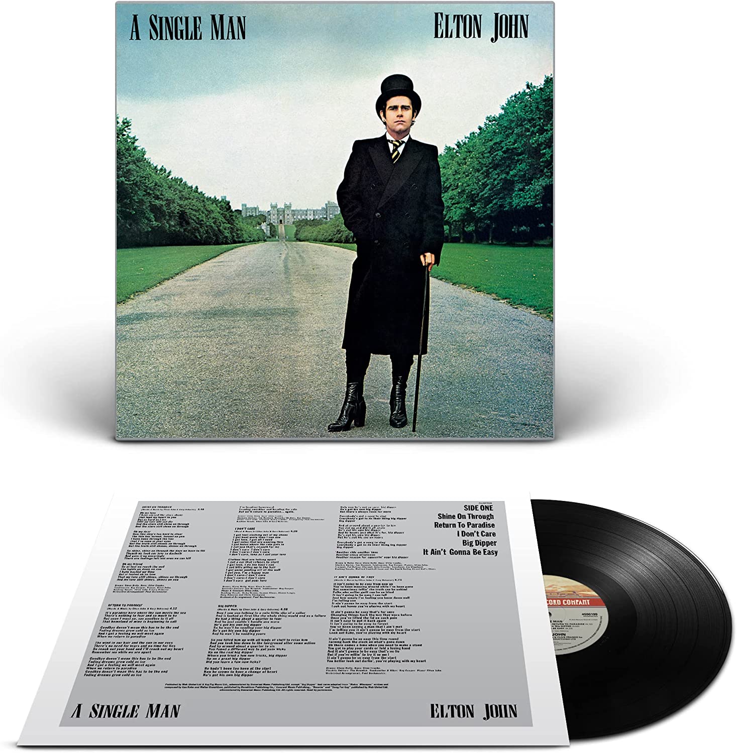 Elton John stands on a driveway, wearing a black coat and top hat, holding a cane.