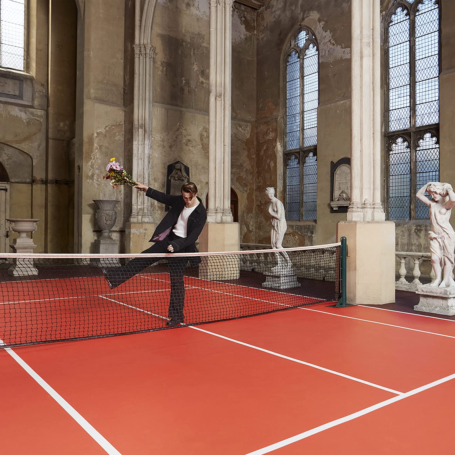 The alt text for the image is: "Man in suit plays tennis in a church with red court."