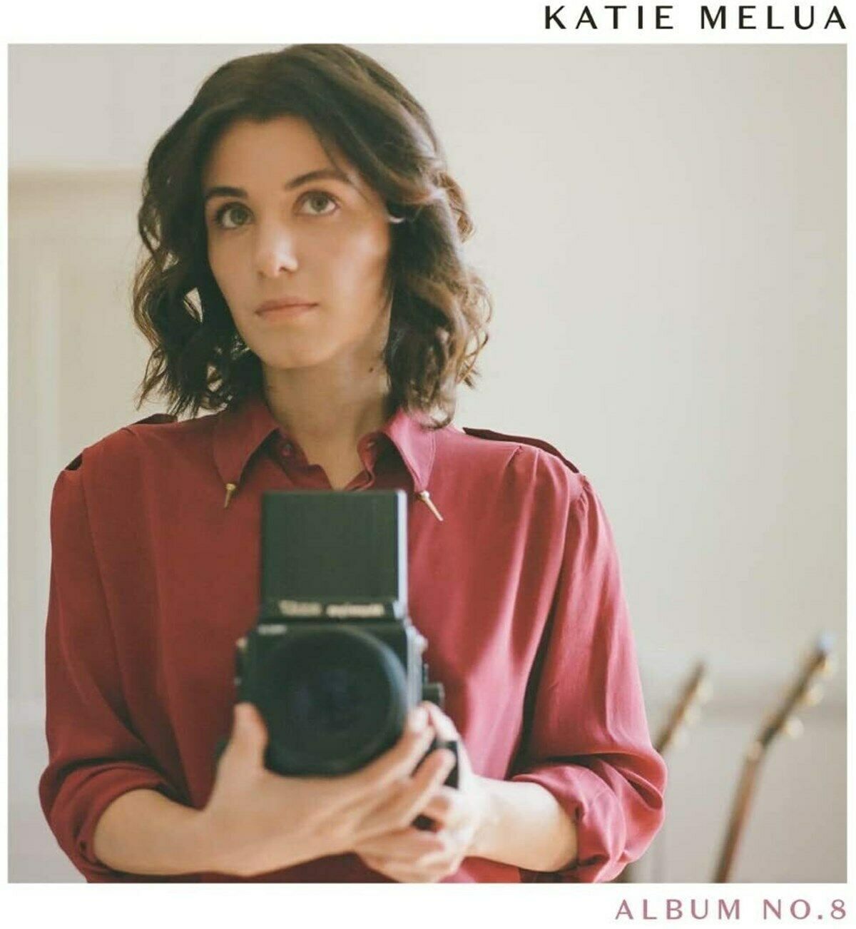 Katie Melua holding a camera, wearing a red shirt, on a white album cover.