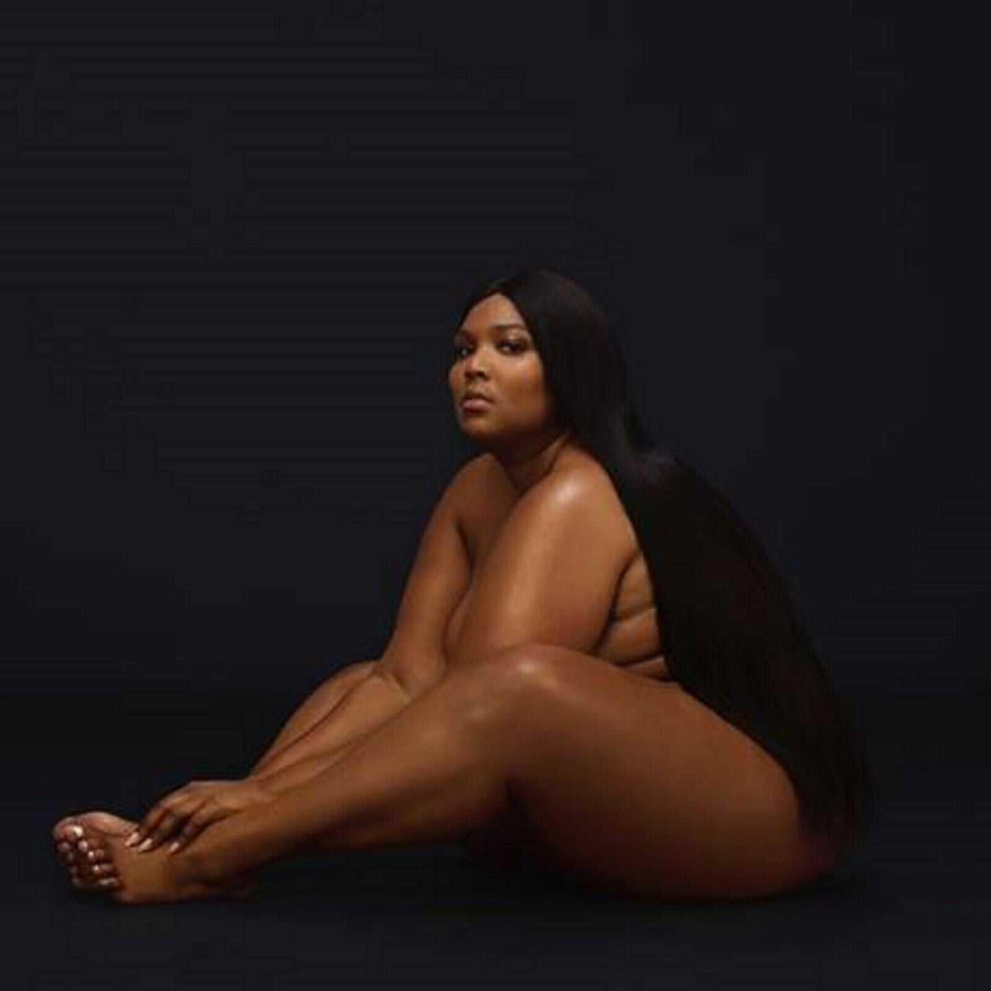 Nude woman with long black hair sits on a dark background.